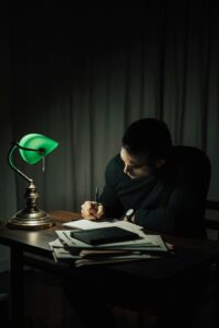 About iam icon advertising and marketing Concentrated male taking notes in notepad while sitting at desktop with documents in dark room with table lamp during paperwork at home